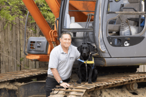 A man kneels next to a black dog wearing a vest, both on the tracks of an excavator.